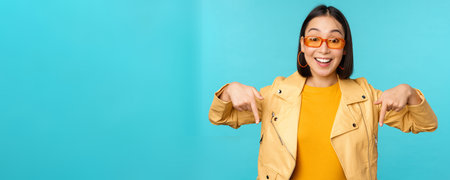 Portrait of stylish asian girl, wears sunglasses, smiles and points fingers down, shows...