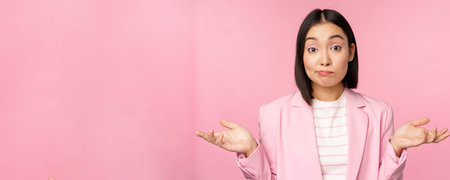 Portrait of confused asian businesswoman shrugging shoulders, looking clueless and puzzled, dont know, cant say, standing over pink background in office suitの写真素材