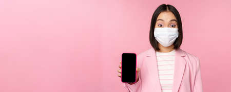 Portrait of smiling korean saleswoman in medical face mask, business suit, showing smartphone screen, standing over pink backgroundの写真素材