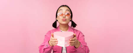 Cute asian girl giving you gift in heart shaped box, kissing and smiling, concept of holiday and celebration, standing over pink backgroundの写真素材