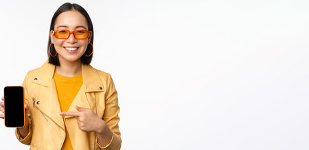 Portrait of stylish korean girl in sunglasses, smiling, pointing finger at smartphone screen,...