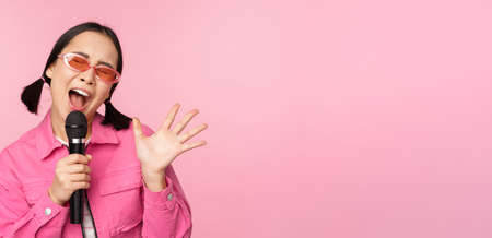 Happy beautiful asian girl singing with mic, using microphone, enjoying karaoke, posing against pink studio backgroundの写真素材