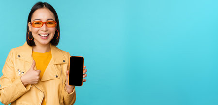 Young stylish chinese woman showing mobile phone screen and thumbs up, smartphone app, standing over...