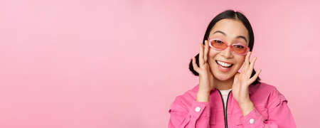 Cute modern japanese girl in sunglasses, smiling and looking happy, posing against pink background in stylish clothingの写真素材