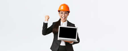 Successful winning businesswoman, construction manager or engineer at factory showing profit chart, diagram at laptop screen, fist pump in rejoice, standing pleased over white backgroundの写真素材