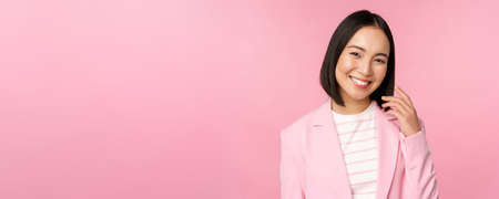 Young professionals. Smiling asian businesswoman, saleswoman in suit looking confident at camera, posing against pink backgroundの写真素材