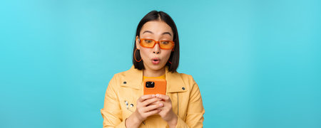 Portrait of asian woman in sunglasses, looking at mobile phone with surprised face expression,...