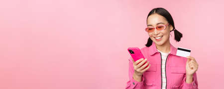 Online shopping. Smiling asian girl shopper, holding smartphone and credit card, paying in mobile app, standing over pink backgroundの写真素材