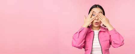 Portrait of asian girl peeks with excitement through fingers, covers eyes, seeing surprise, standing over pink backgroundの写真素材