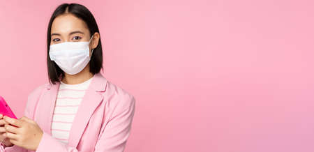 Asian businesswoman in medical face mask using mobile phone. Japenese saleswoman, corporate lady in suit, holding smartphone, standing over pink backgroundの写真素材
