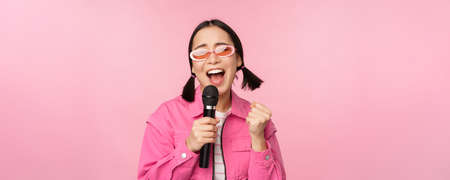 Happy beautiful asian girl singing with mic, using microphone, enjoying karaoke, posing against pink studio backgroundの写真素材