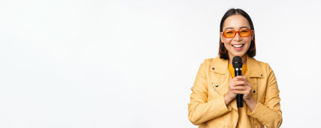 Stylish asian girl in sunglasses, singing songs with microphone, holding mic and dancing at karaoke,...