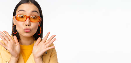 Stylish young asian woman in trendy sunglasses, pucker lips silly, looking surprised, impressed reaction, standing over white backgroundの写真素材
