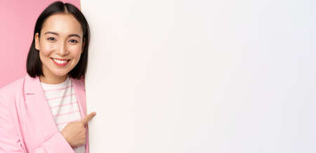 Portrait of happy smiling japanese corporate woman pointing finger at board, showing smth on empty space, standing in suit over pink backgroundの写真素材