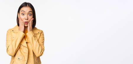 Image of asian woman looking surprised, amazed reaction, shocked face, standing over white backgroundの写真素材