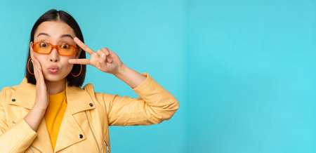Portrait of attractive korean woman in sunglasses, showing peace v-sign near eyes, pucker lips,...