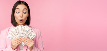 Finance, microcredit and people concept. Happy smiling asian businesswoman showing dollars money, standing in suit against pink backgroundの写真素材