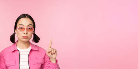 Close up portrait of asian stylish girl, looking skeptical, pointing finger up with unamused face, standing over pink backgroundの写真素材