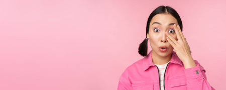 Close up portrait of young asian girl looking surprised, express amazement and wonder, peeking...