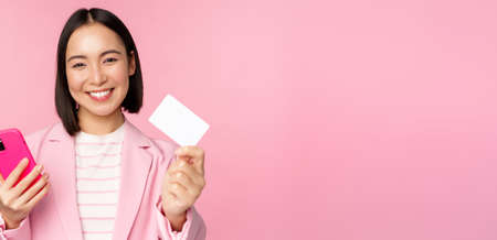 Image of smiling happy asian businesswoman showing credit card, paying online on smartphone application, order with mobile phon, standing against pink backgroundの写真素材