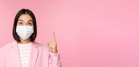 Close up portrait of businesswoman, asian lady in office suit and medical face mask, looking and pointing up, showing company logo or banner on top, pink backgroundの写真素材