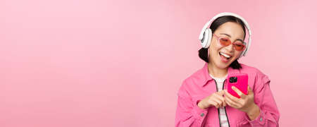 Stylish asian girl dancing with smartphone, listening music in headphones on mobile phone app, smiling and laughing, posing against pink backgroundの写真素材