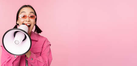 Attention, announcement concept. Enthusiastic asian girl shouting in megaphone, advertising with speaker, recruiting, standing over pink backgroundの写真素材