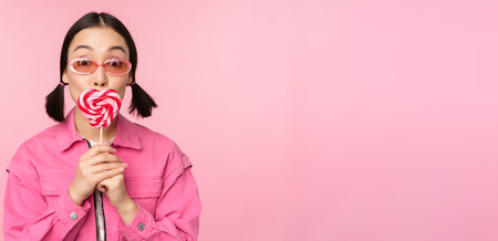 Stylish korean girl licking lolipop, eating candy and smiling, standing in sunglasses against pink backgroundの写真素材