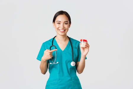 Healthcare workers, preventing virus, quarantine campaign concept. Smiling pretty asian female physician, nurse in scrubs show vitamins and pointing at pills, recommend medication, white backgroundの写真素材