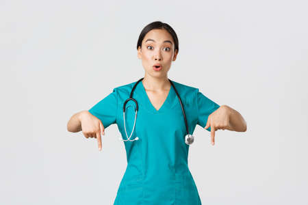 Covid-19, healthcare workers, pandemic concept. Amazed and impressed asian female nurse, doctor in scrubs pointing fingers down and looking questioned, interested in new promo, white backgroundの写真素材
