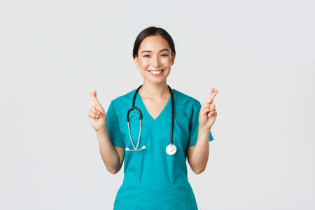 Covid-19, healthcare workers, pandemic concept. Optimistic smiling asian female doctor, physician in scrubs having faith, cross fingers good luck, making wish, hope for better, white backgroundの写真素材
