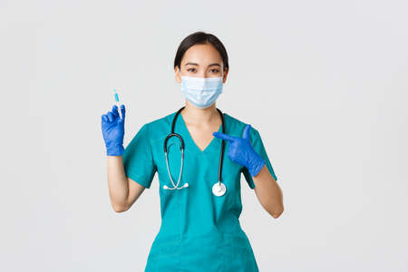 Covid-19, coronavirus disease, healthcare workers concept. Confident friendly-looking asian female doctor, physician in medical mask and scrubs pointing finger at syringe with vaccineの写真素材