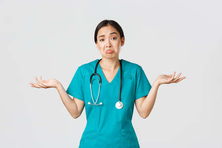 Covid-19, healthcare workers, pandemic concept. Portrait of clueless asian female nurse, woman doctor shrugging and spread hands sideways unaware, dont know, cant help, white backgroundの写真素材