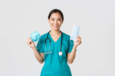 Covid-19, coronavirus disease, healthcare workers concept. Smiling pretty asian female nurse, physician in scrubs showing respirator and medical mask, suggest patients ways of virus protectionの写真素材