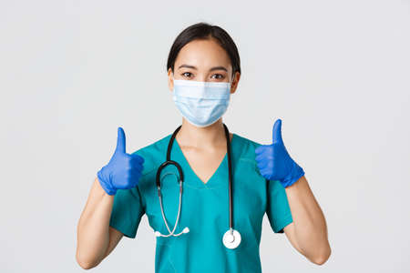 Covid-19, coronavirus disease, healthcare workers concept. Confident smiling asian doctor, nurse in medical mask and rubber gloves, show thumbs-up in approval, provide service, wite backgroundの写真素材