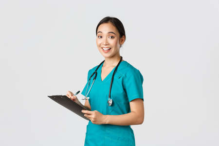 Healthcare workers, preventing virus, quarantine campaign concept. Smiling cute asian female nurse, intern in scrubs, writing on clipboard, fill-in papers, looking a camera pleased, white backgroundの写真素材