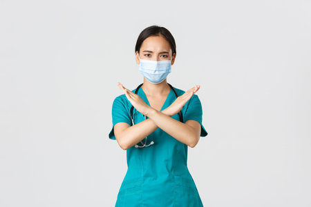 Covid-19, coronavirus disease, healthcare workers concept. Serious displeased, frowning asian doctor in scrubs show cross gesture in disapproval, stop someone, prohibit action, white backgroundの写真素材