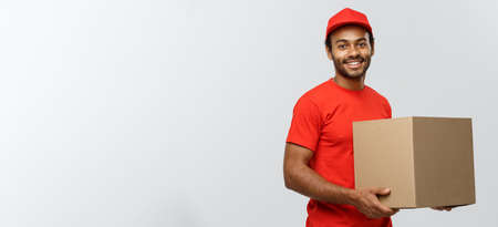 Delivery Concept - Portrait of Happy African American delivery man in red cloth holding a box package. Isolated on Grey studio Background. Copy Space.の写真素材