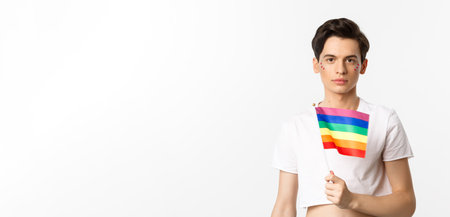 Pride and lgbtq concept. Waist up shot of attractive anrogynous man holding rainbow flag, having glitter on face and looking at camera, standing over white backgroundの写真素材