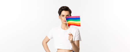 Lgbtq community. Attractive queer man with flitter on face, waving pride rainbow flag and looking at camera, standing in crop top against white backgroundの写真素材