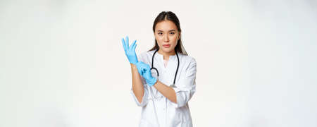 Confident female nurse, physician put on rubber medical gloves for patient clinical examination, standing serious in healthcare worker uniform, white backgroundの写真素材