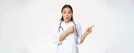 Surprised female doctor say wow, pointing right and looking amazed with discounts in clinic, showing way, standing in medical uniform against white backgroundの写真素材