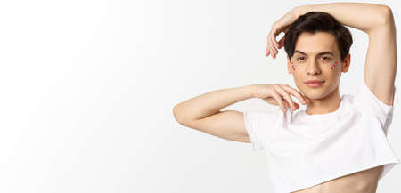 People, lgbtq community and lifestyle concept. Close-up of sassy gay man with lip gloss and glitter under eyes, posing for camera with pleased smile, wearing crop top, white backgroundの写真素材