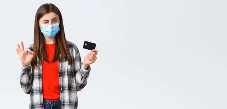 Coronavirus outbreak, working from home, online shopping and contactless payment concept. Pleased young female bank customer in medical mask, show okay sign and credit card, winkの写真素材