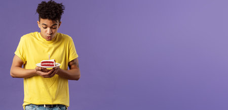Portrait of cute excited young guy with dreads and tattoos, making wish on birthday, celebrating party, blowing candle on b-day cake, standing purple background dreamingの写真素材
