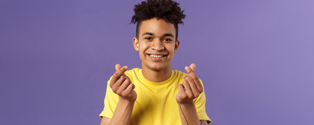 Close-up portrait of lovely cute young man with afro haircut, showing korean heart finger signs and smiling, express his love and sympathy towards person, stand purple backgroundの写真素材