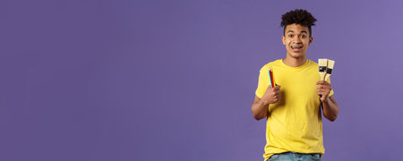 Portrait of upbeat young creative man, artist starting art courses, want to learn how to draw, holding colored pencils and brushes, standing enthusiastic over purple backgroundの写真素材