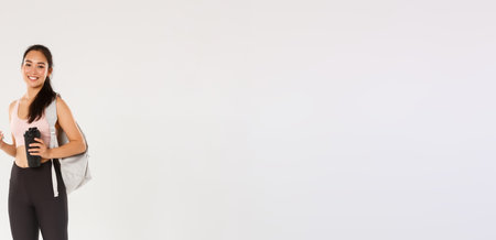 Full length of smiling healthy, slim asian girl going fitness training, female athelte carry backpack with workout equipment and water bottle, using mobile phone sports application, white backgroundの写真素材