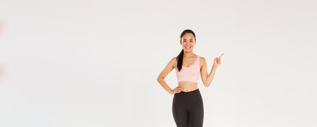 Sport, gym and healthy body concept. Full length of smiling pretty asian fitness girl, female athlete in active wear pointing finger left, gym coach showing training equipment, white backgroundの写真素材