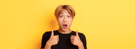 Close-up of impressed and excited handsome asian guy showing thumbs-up and looking amazed, standing...